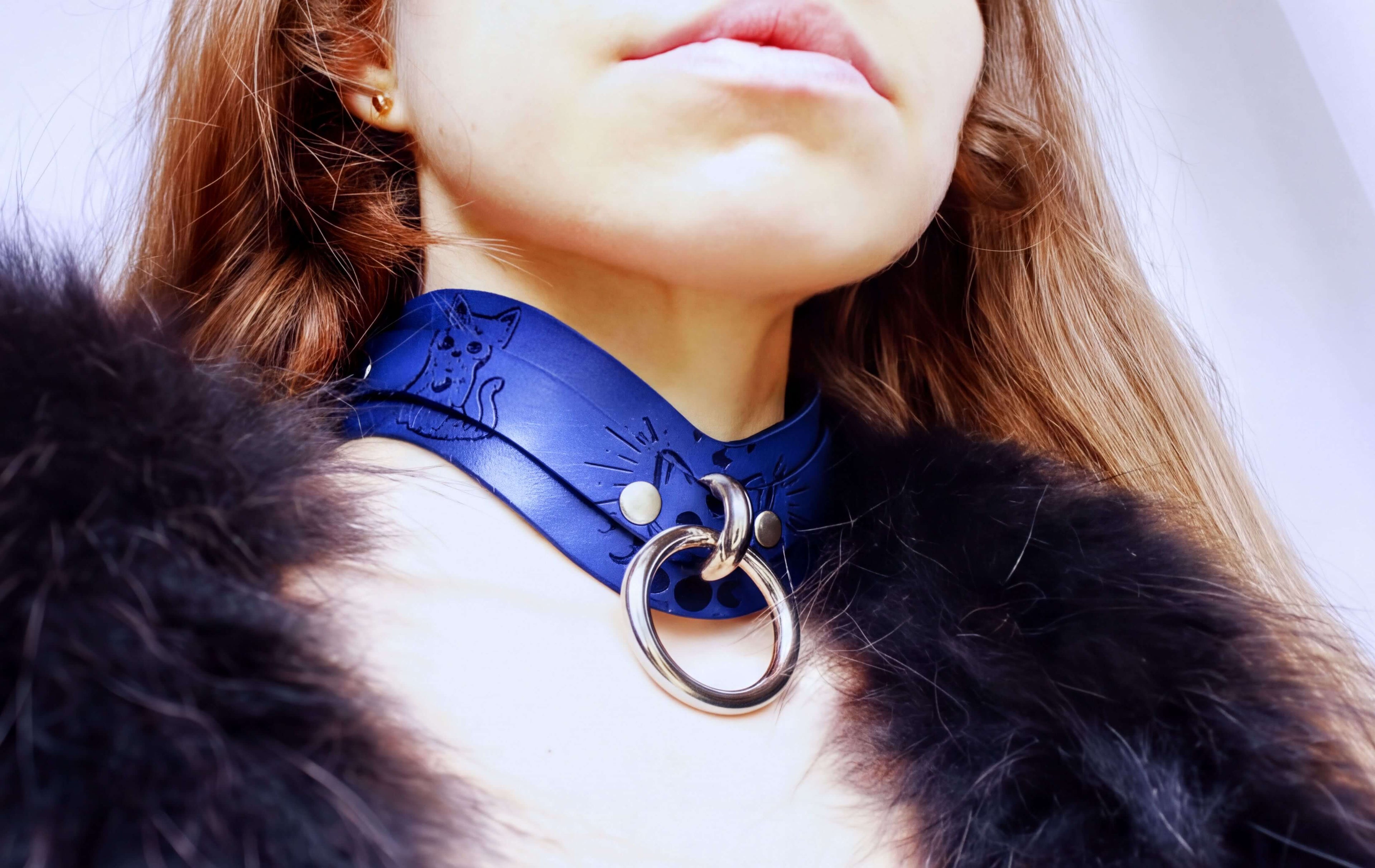 A close-up of a woman's neck and chest area, featuring a blue choker necklace with a metal ring and a fur collar.