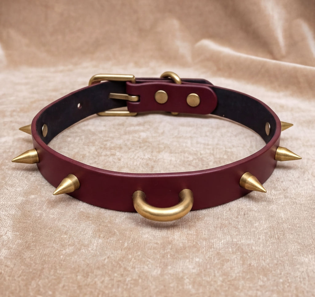 A burgundy leather collar with gold spikes protruding from it, resting on a beige fabric background.