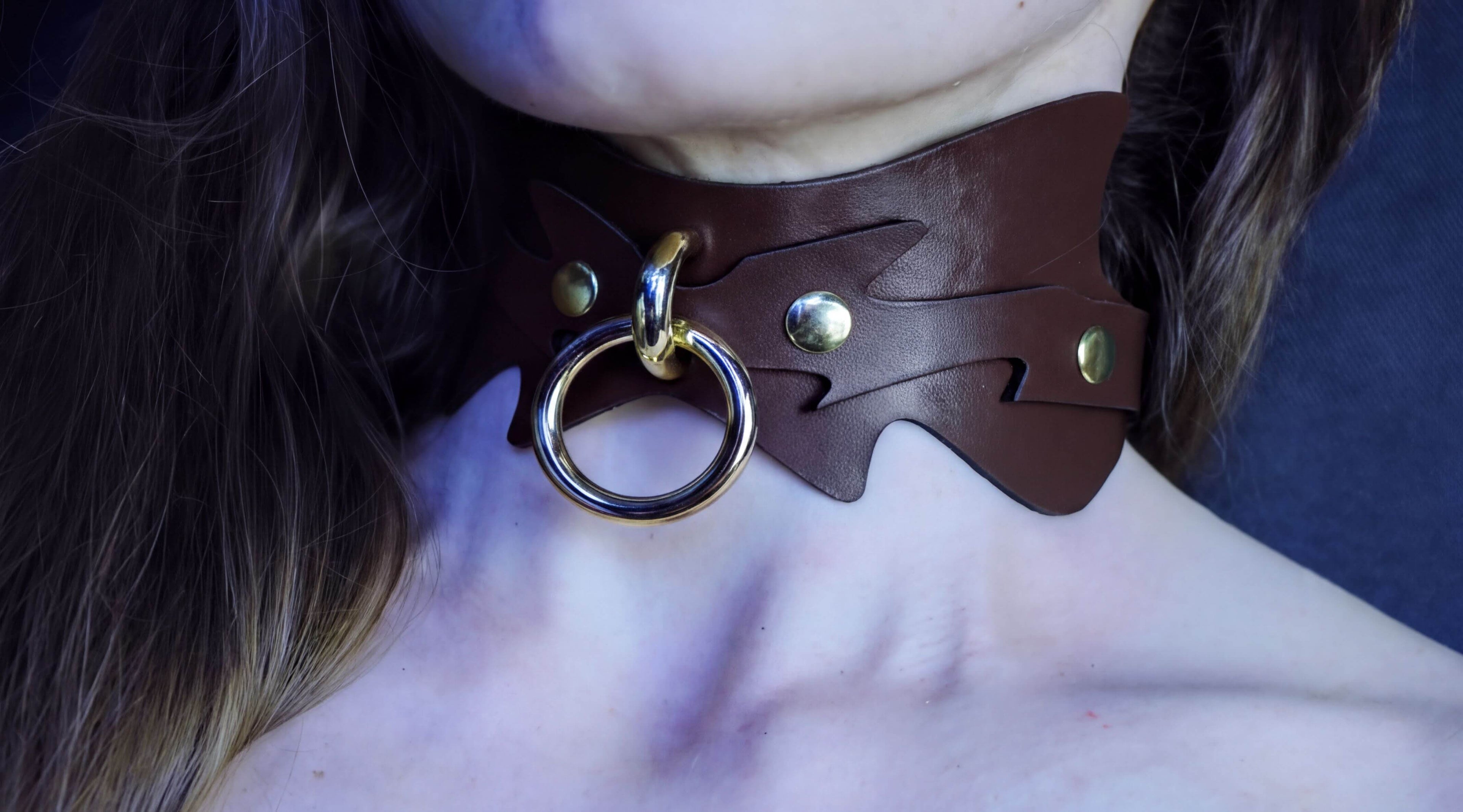 A close-up of a person's neck wearing a brown leather choker necklace with a metal ring and two gold-colored buttons.