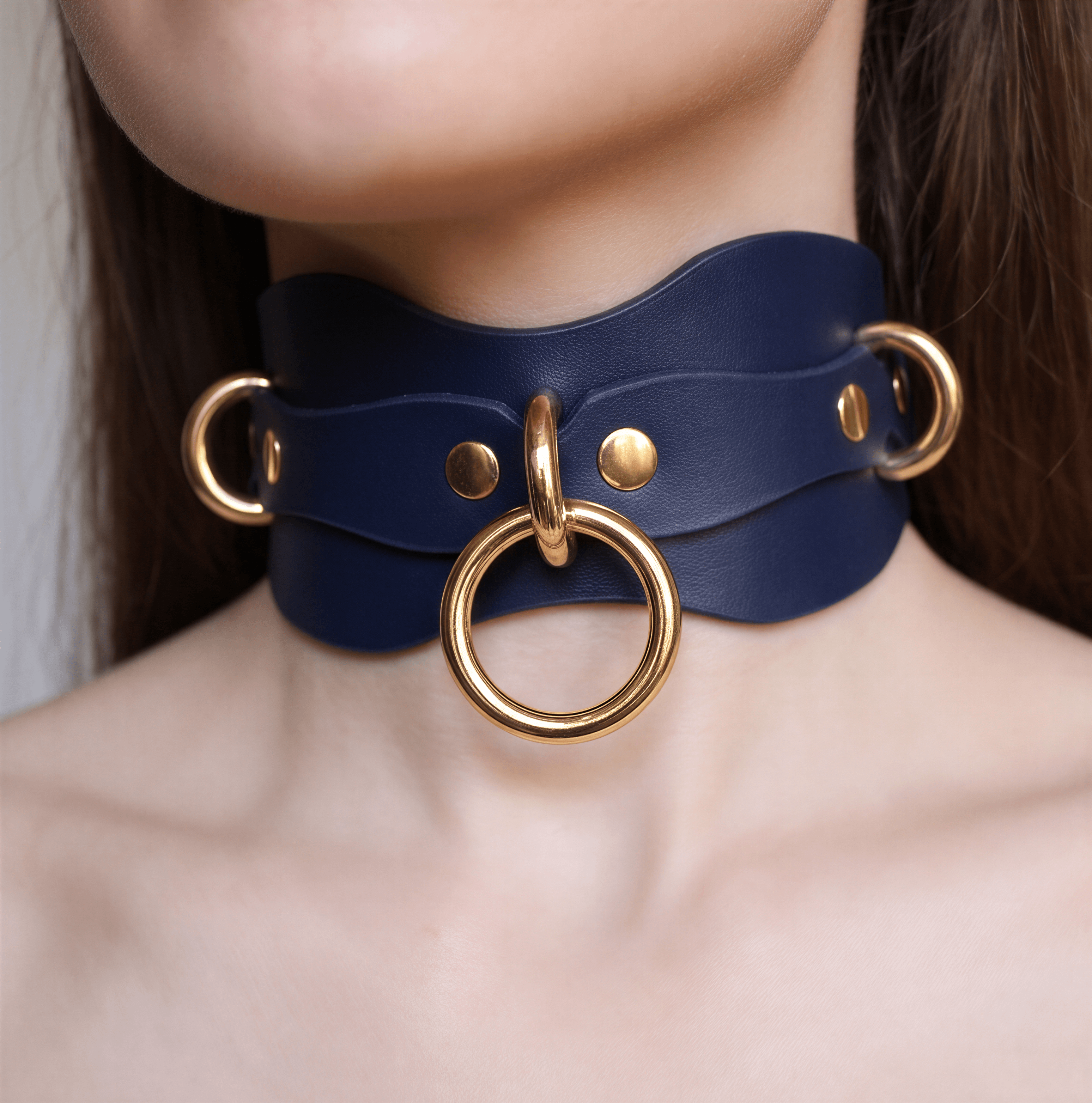 A close-up of a person's neck wearing a blue leather choker necklace with gold rings and a large gold ring on the front.