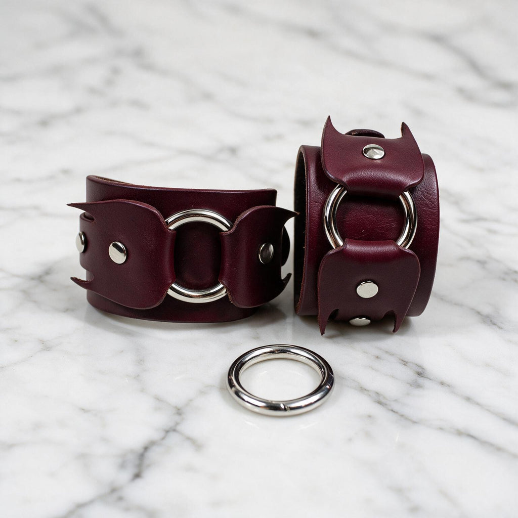 A pair of burgundy leather cuffs with metal rings and a ring attachment on a marble surface.