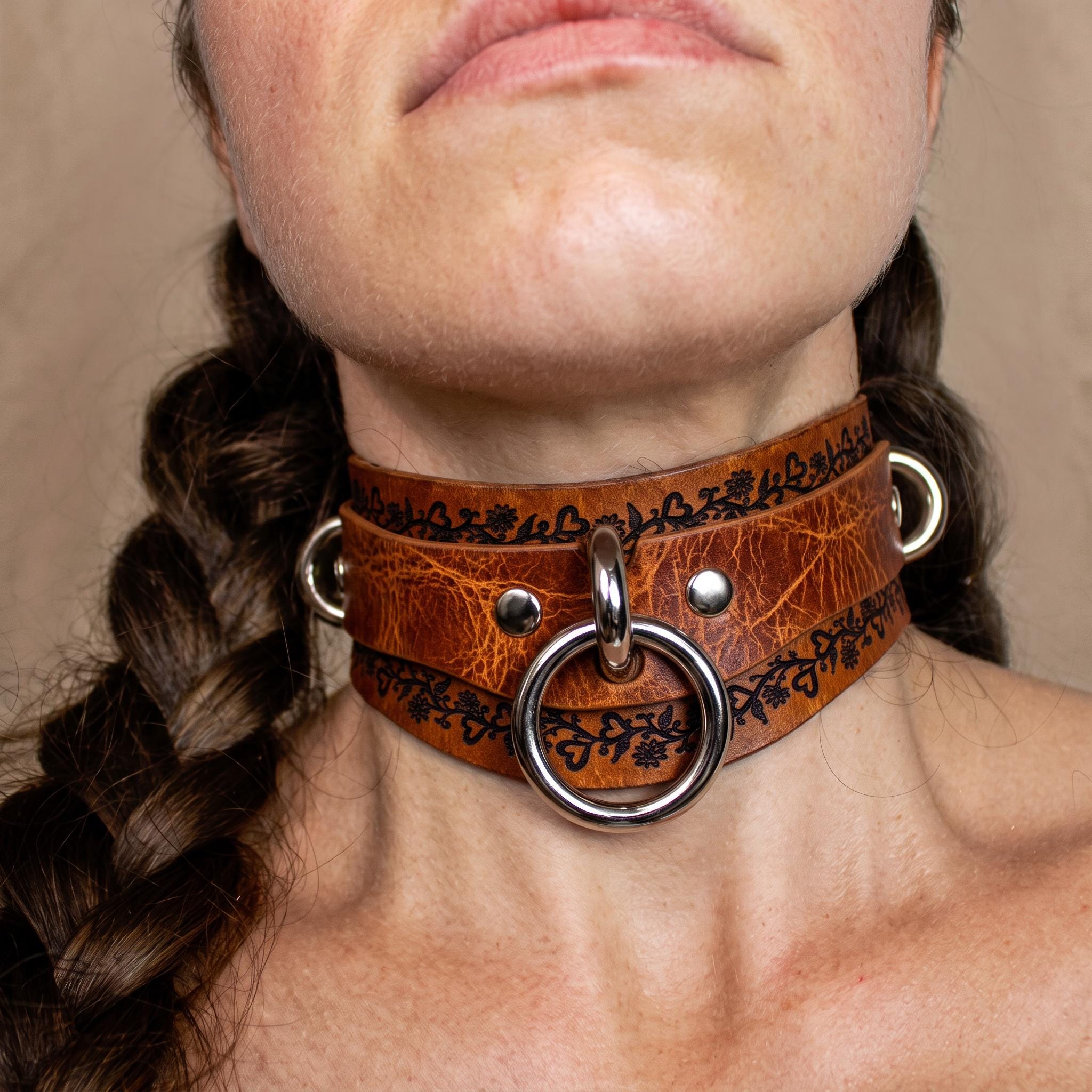 A close-up of a woman's neck and chin wearing a leather choker necklace with metal rings and a metal ring.