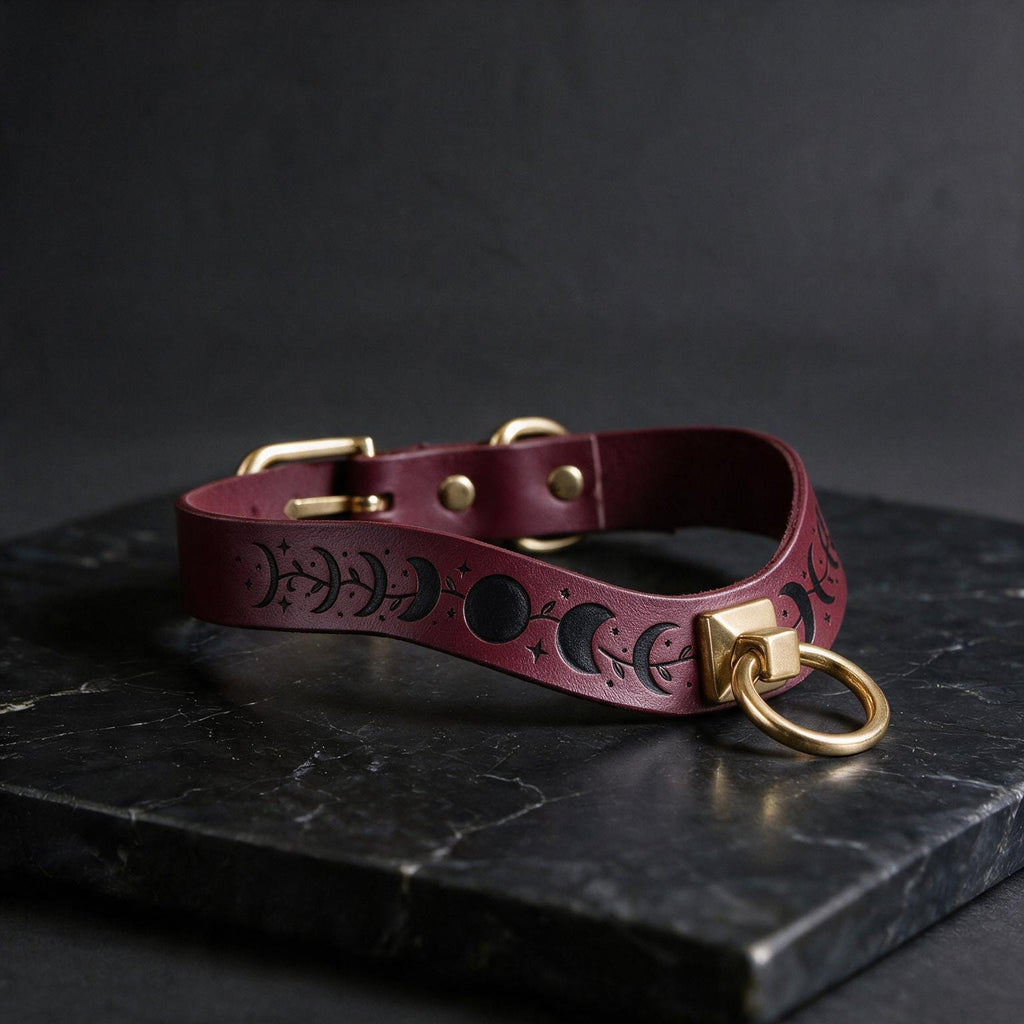 A burgundy leather collar with a moon and star motif, featuring a gold-colored metal ring and buckle, is displayed on a black marble surface.