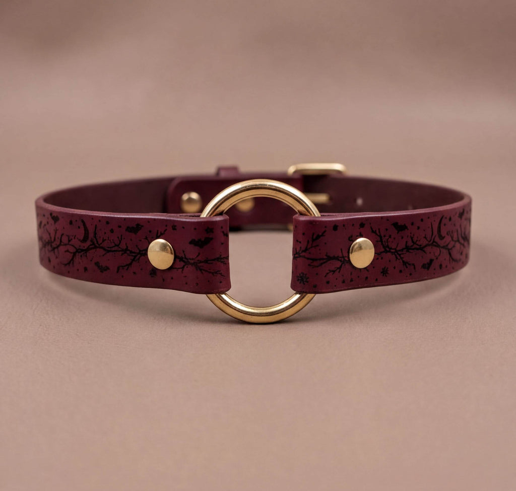 A burgundy leather collar with a gold-colored circular buckle and a black leaf design on the strap.