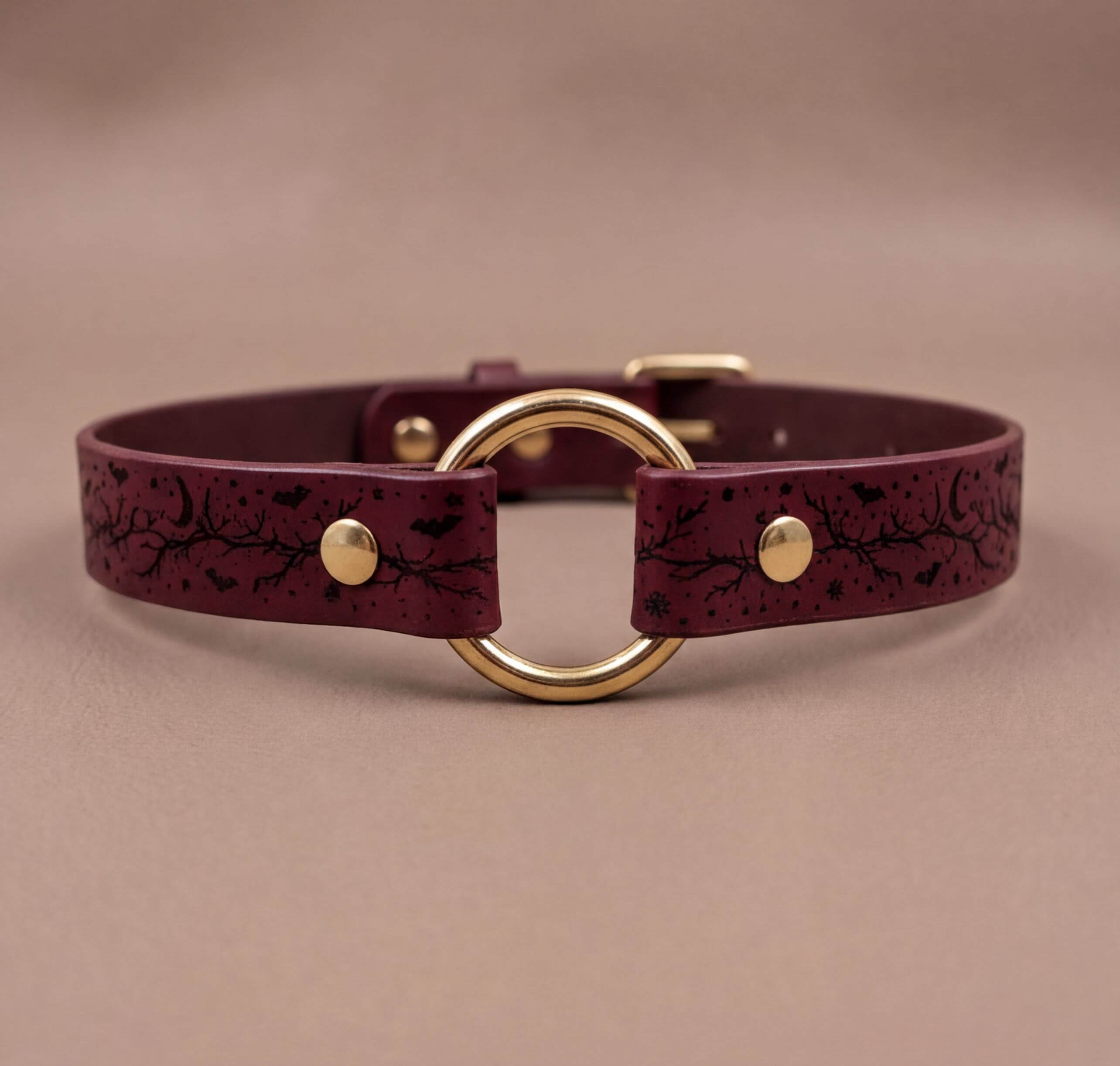 A burgundy leather collar with a gold-colored circular buckle and a black leaf design on the strap.