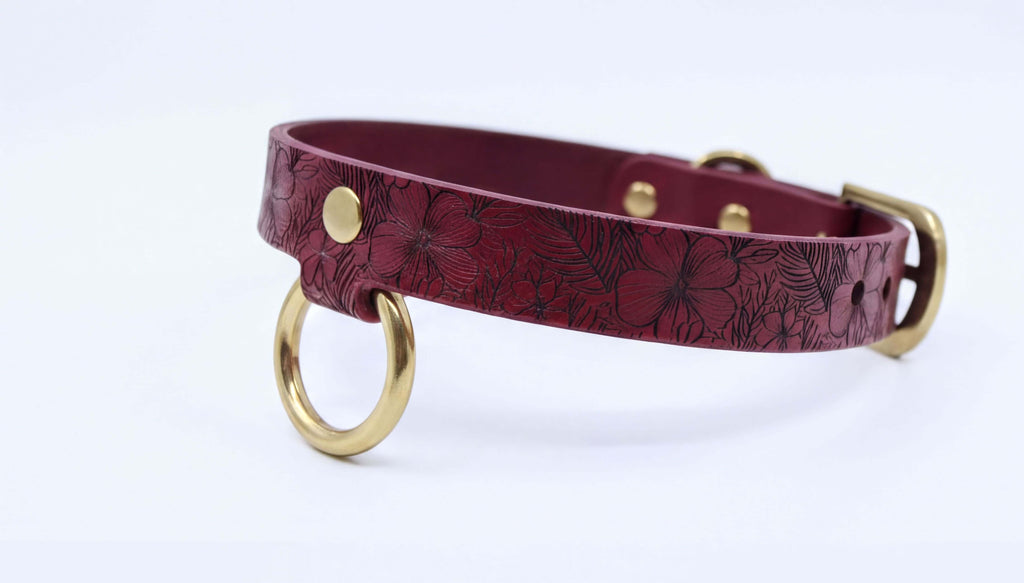 A burgundy leather dog collar with a floral pattern and two gold-colored metal rings.