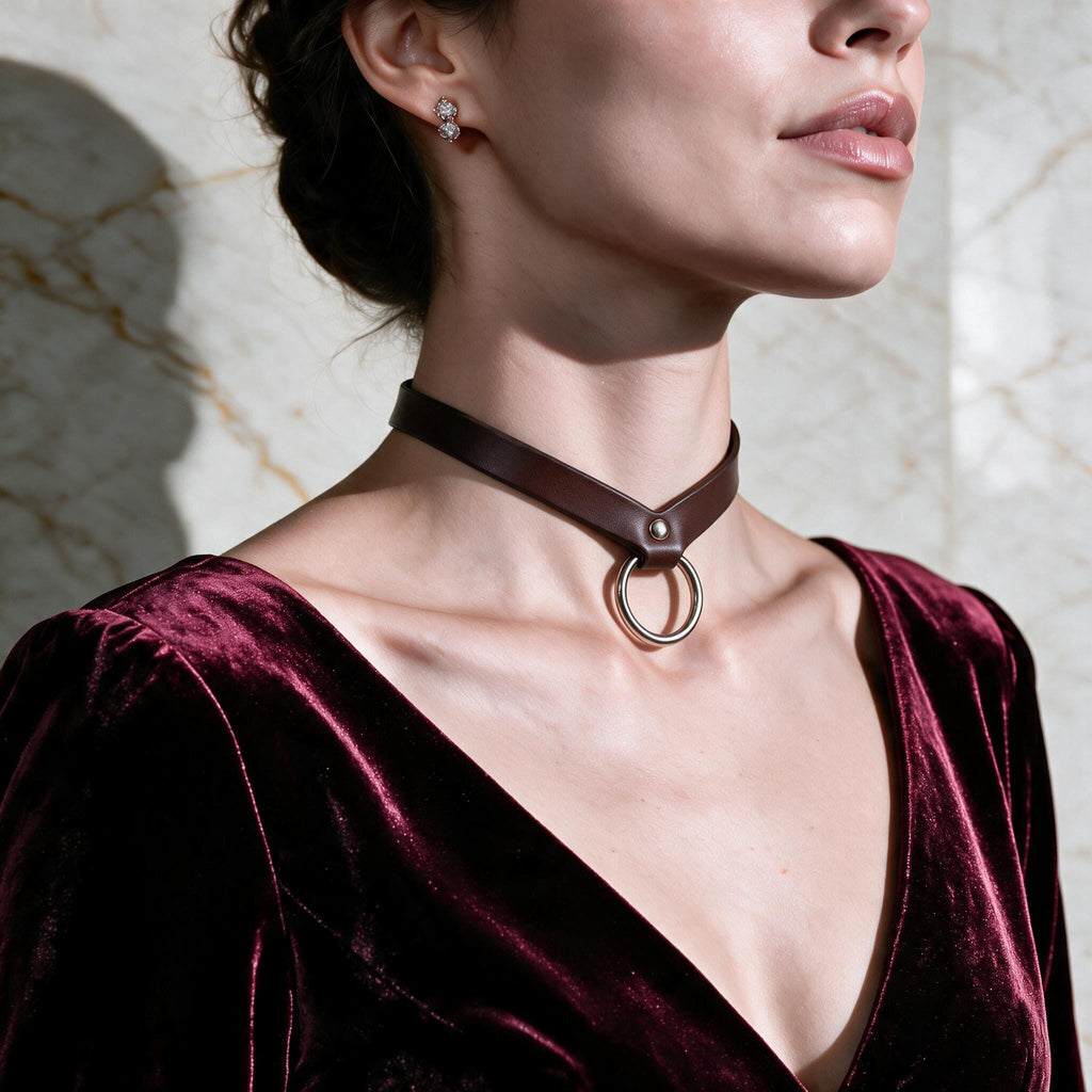 A woman wearing a burgundy velvet dress with a choker necklace and earrings stands against a marble wall.