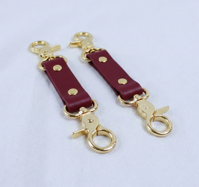 a pair of burgundy leather straps with gold metal clasps and hooks, placed on a white surface.