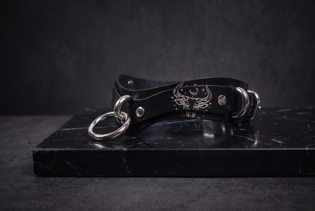 A black leather choker necklace with metal rings and a skull design on the clasp is displayed on a black marble surface.