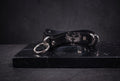 A black leather choker necklace with metal rings and a skull design on the clasp is displayed on a black marble surface.