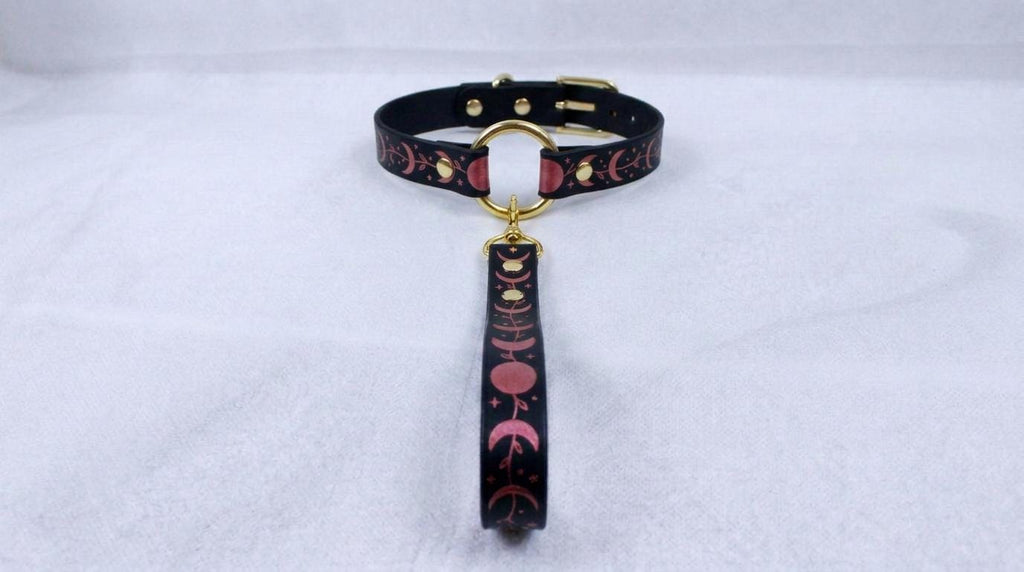 A black collar with a pink and black patterned strap and a gold-colored buckle is displayed on a white surface.