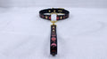 A black collar with a pink and black patterned strap and a gold-colored buckle is displayed on a white surface.