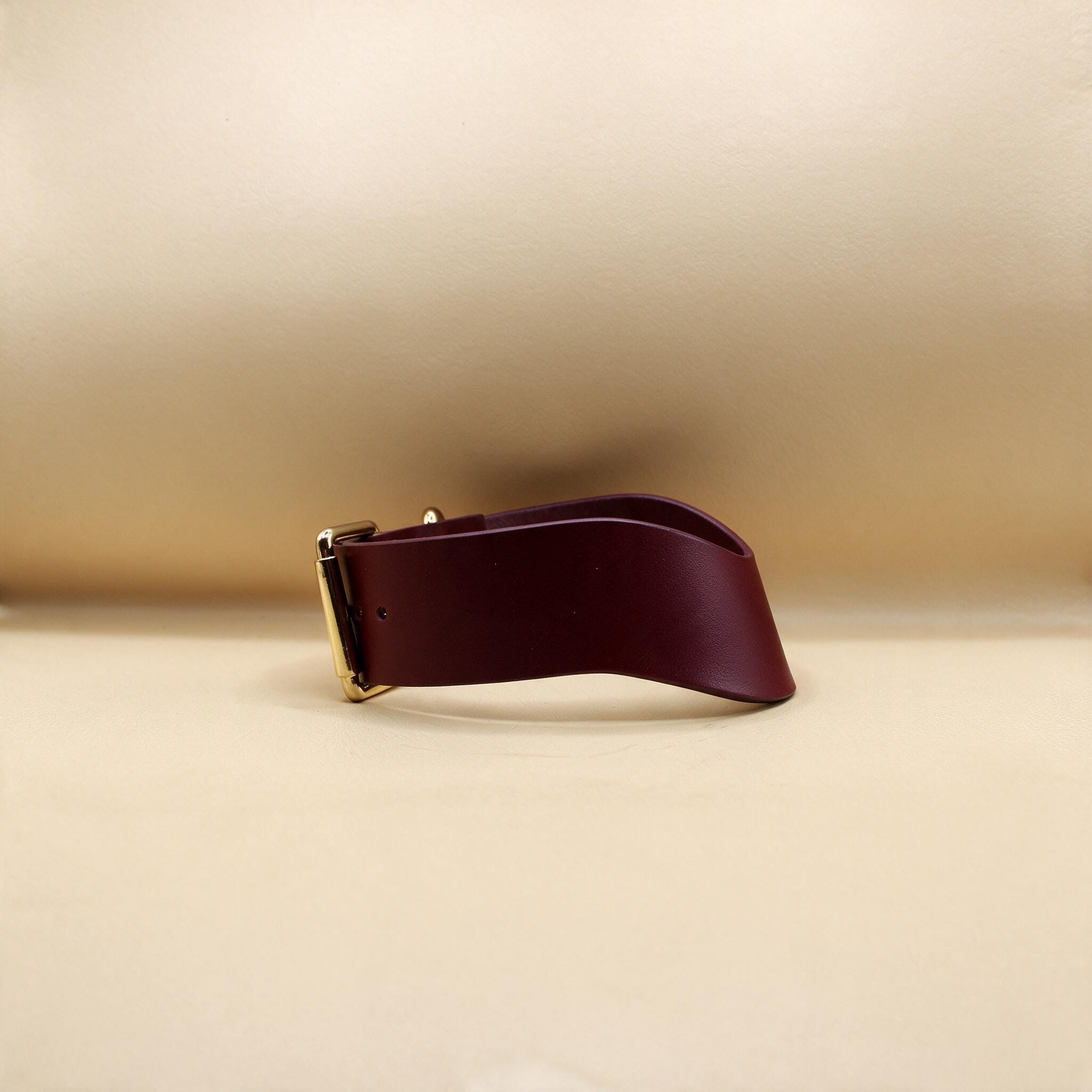 A burgundy leather belt with a gold buckle is shown against a beige background.