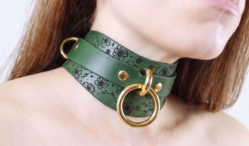 A close-up of a woman's neck wearing a green leather choker necklace with a gold ring and two gold rings on the collar.