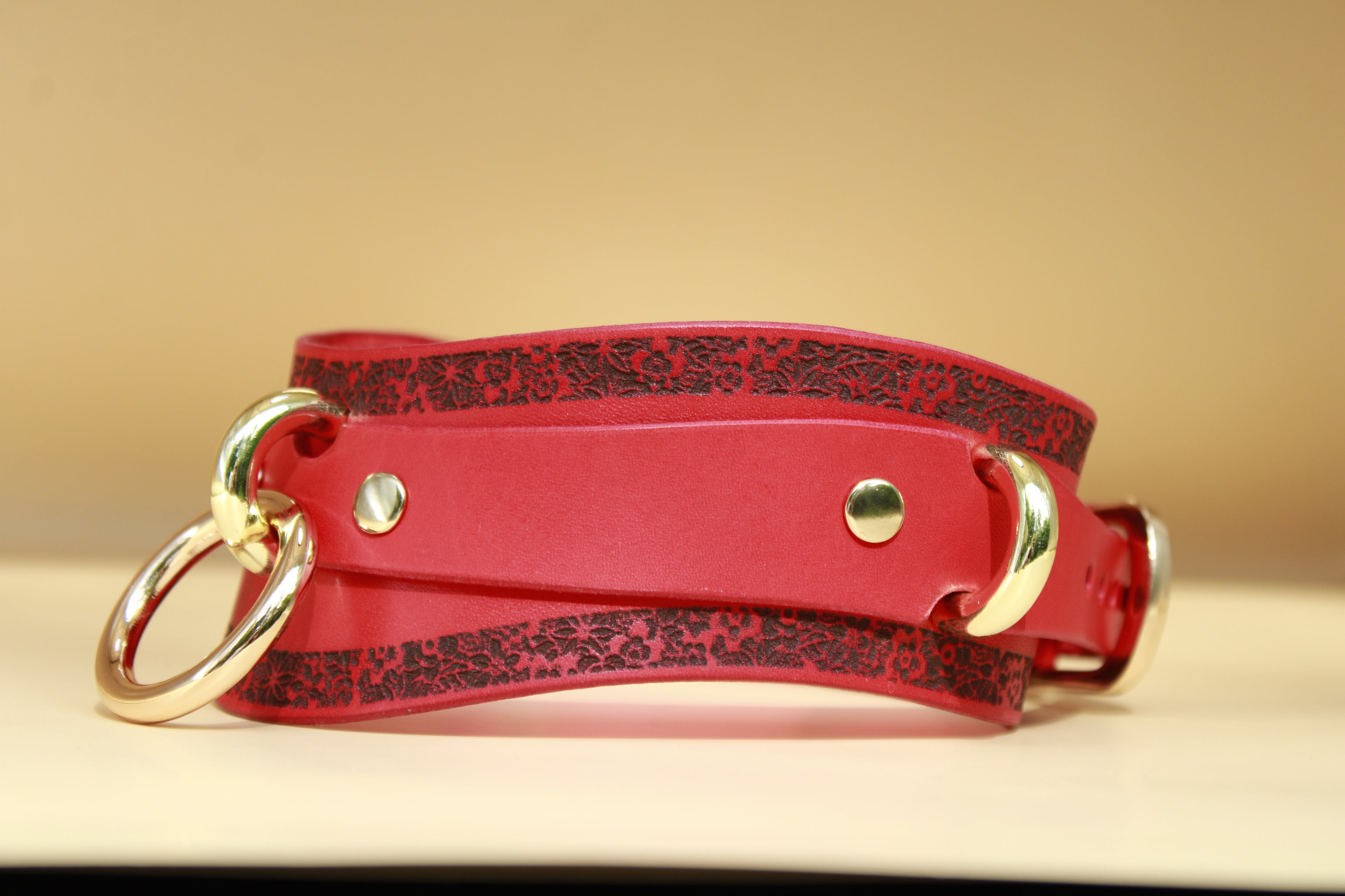 A red leather choker with a floral pattern and two gold-colored buckles is displayed on a light-colored surface.
