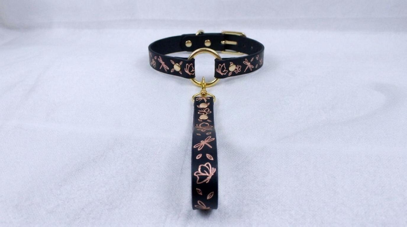 A black collar with a gold-colored buckle and a floral pattern on the strap is displayed against a white background.