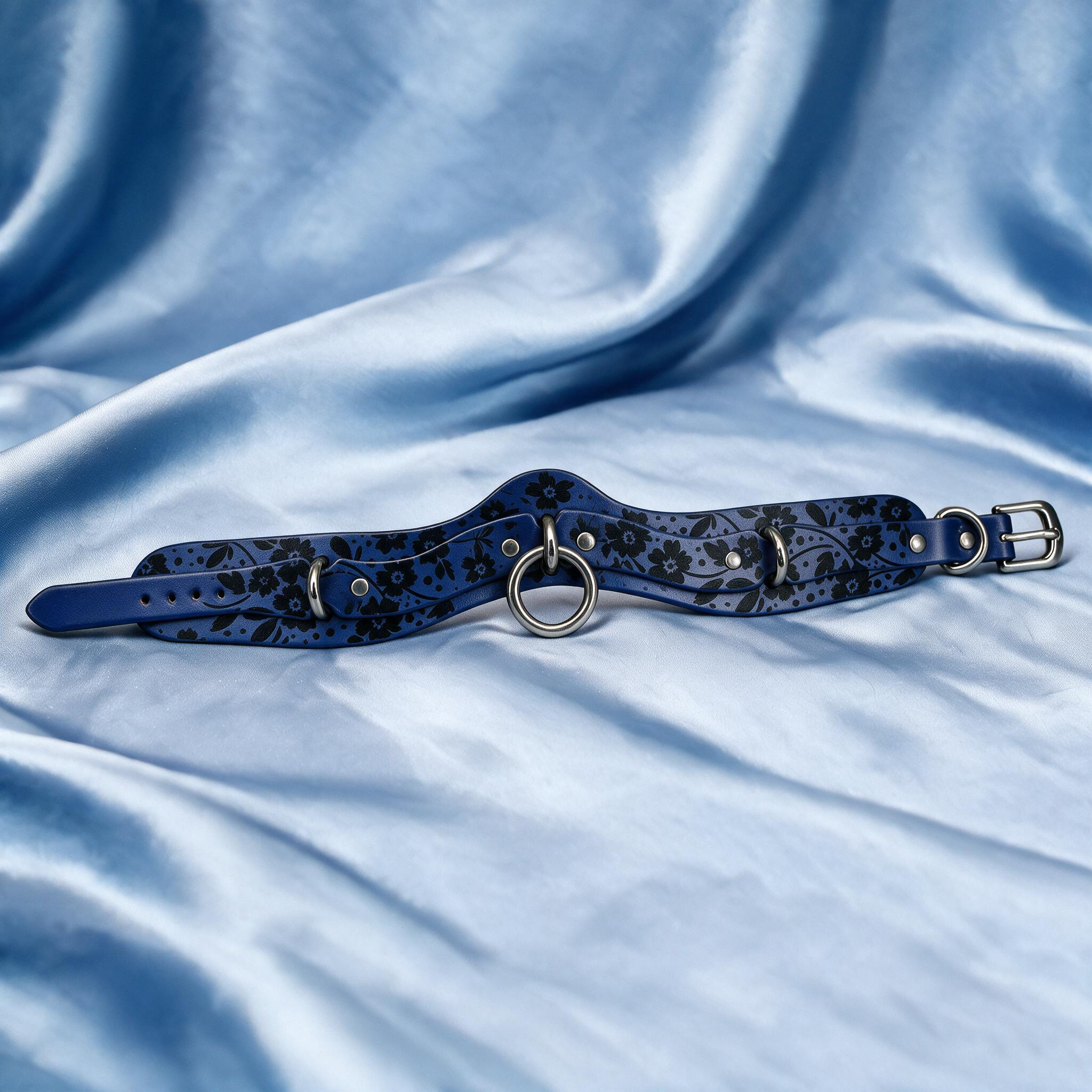 A blue choker necklace with a floral pattern and metal rings is displayed on a blue satin background.