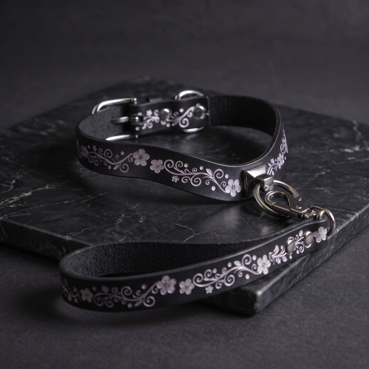 A black leather collar with a floral pattern and a matching leash are displayed on a dark surface.