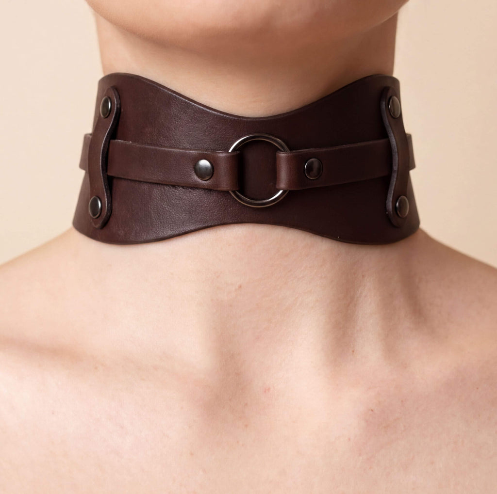 A close-up view of a person's neck and chest area wearing a brown leather choker necklace with metal rings and buckles.