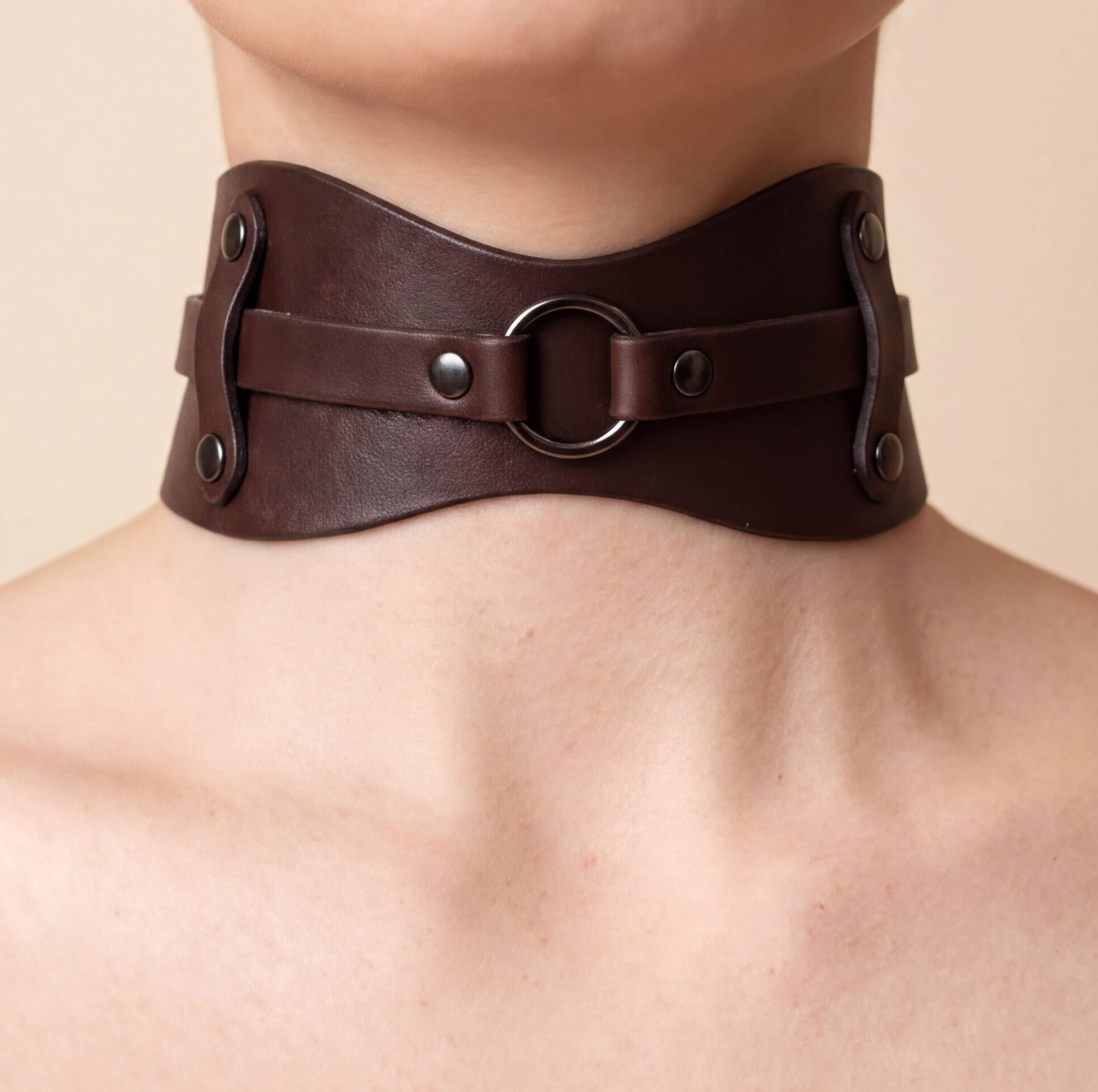 A close-up view of a person's neck and chest area wearing a brown leather choker necklace with metal rings and buckles.