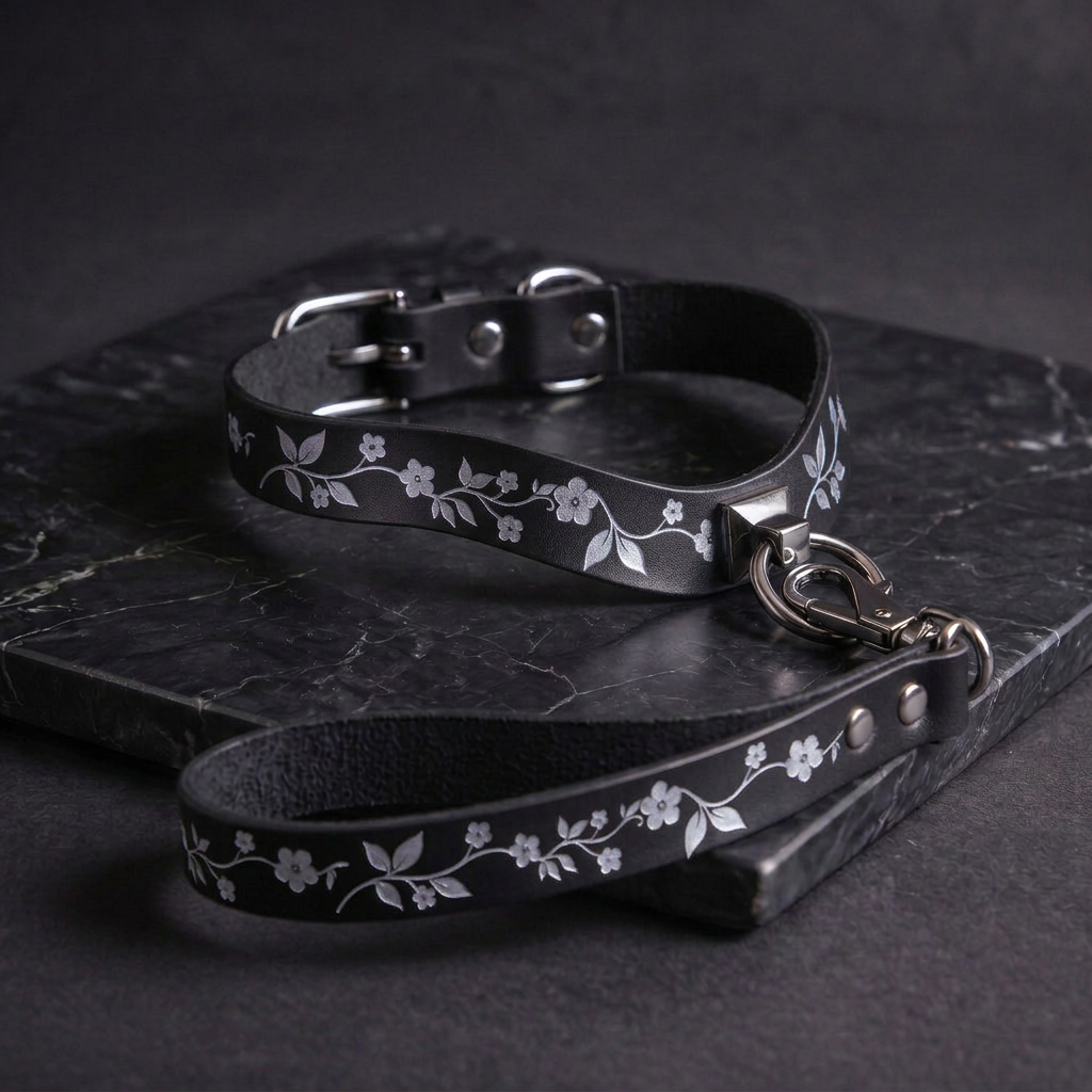 A black leather collar with white floral designs and a matching leash are displayed on a dark surface.