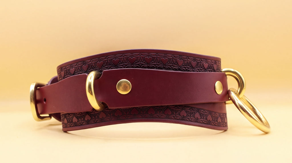 A burgundy leather choker with gold hardware, including two round buckles and two metal rings, is shown against a yellow background.