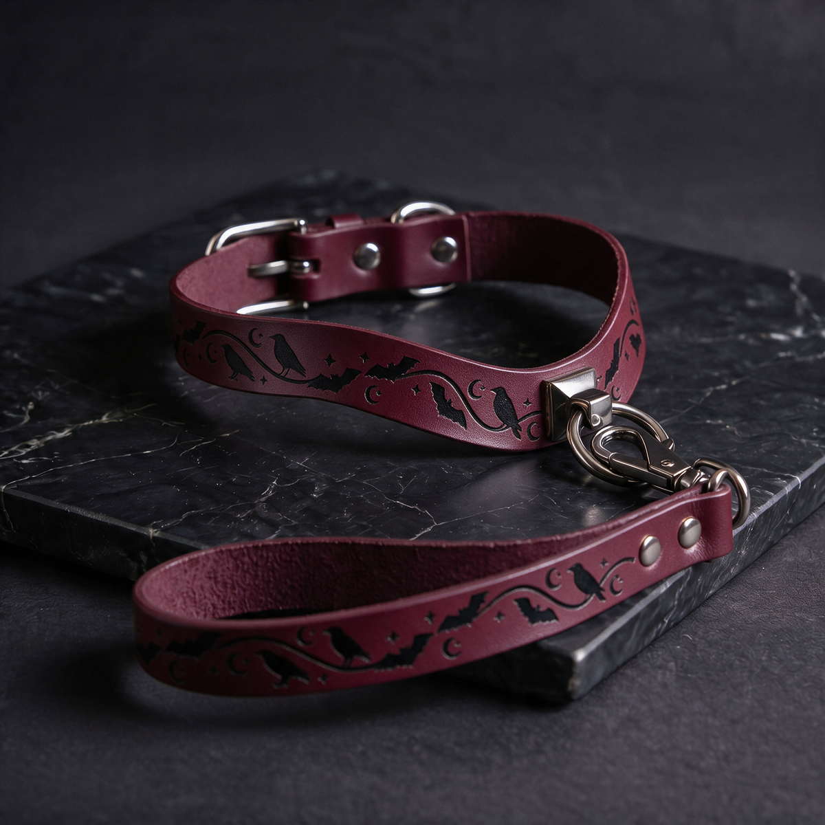 A burgundy leather dog collar with a black bat and moon design, and a matching leash, resting on a dark surface.