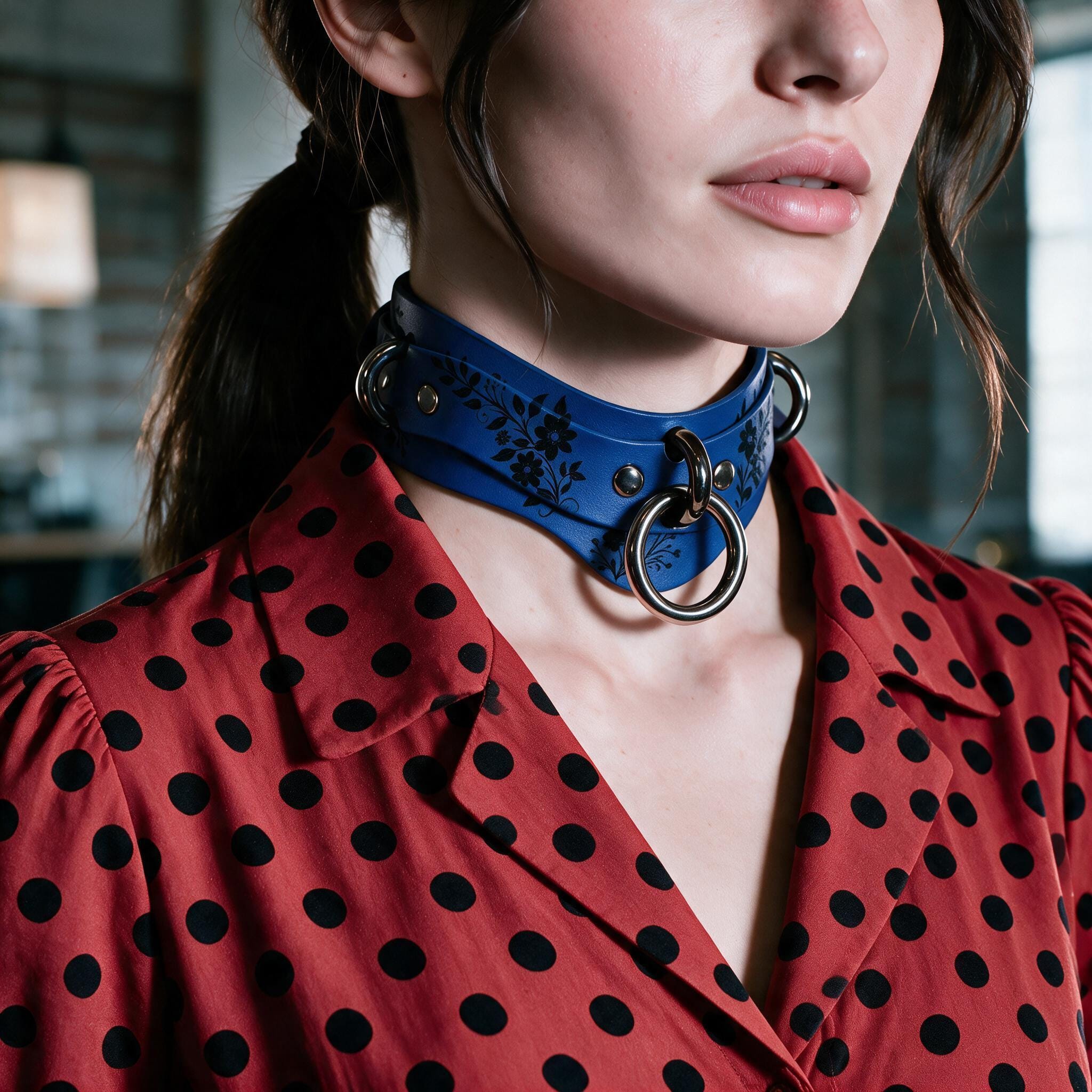 A close-up of a woman wearing a blue choker necklace with metal rings, paired with a red polka-dot blouse.