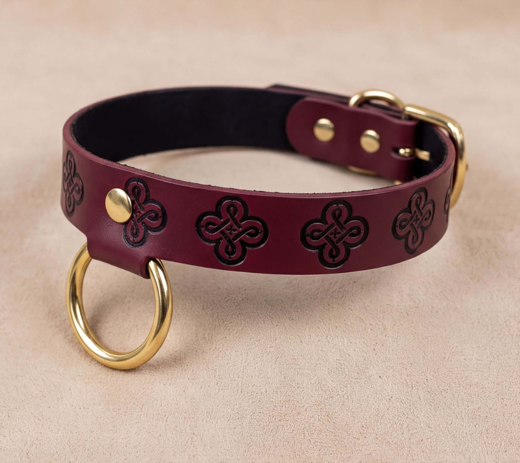 A burgundy leather dog collar with a gold-colored metal ring and a decorative pattern on the leather.