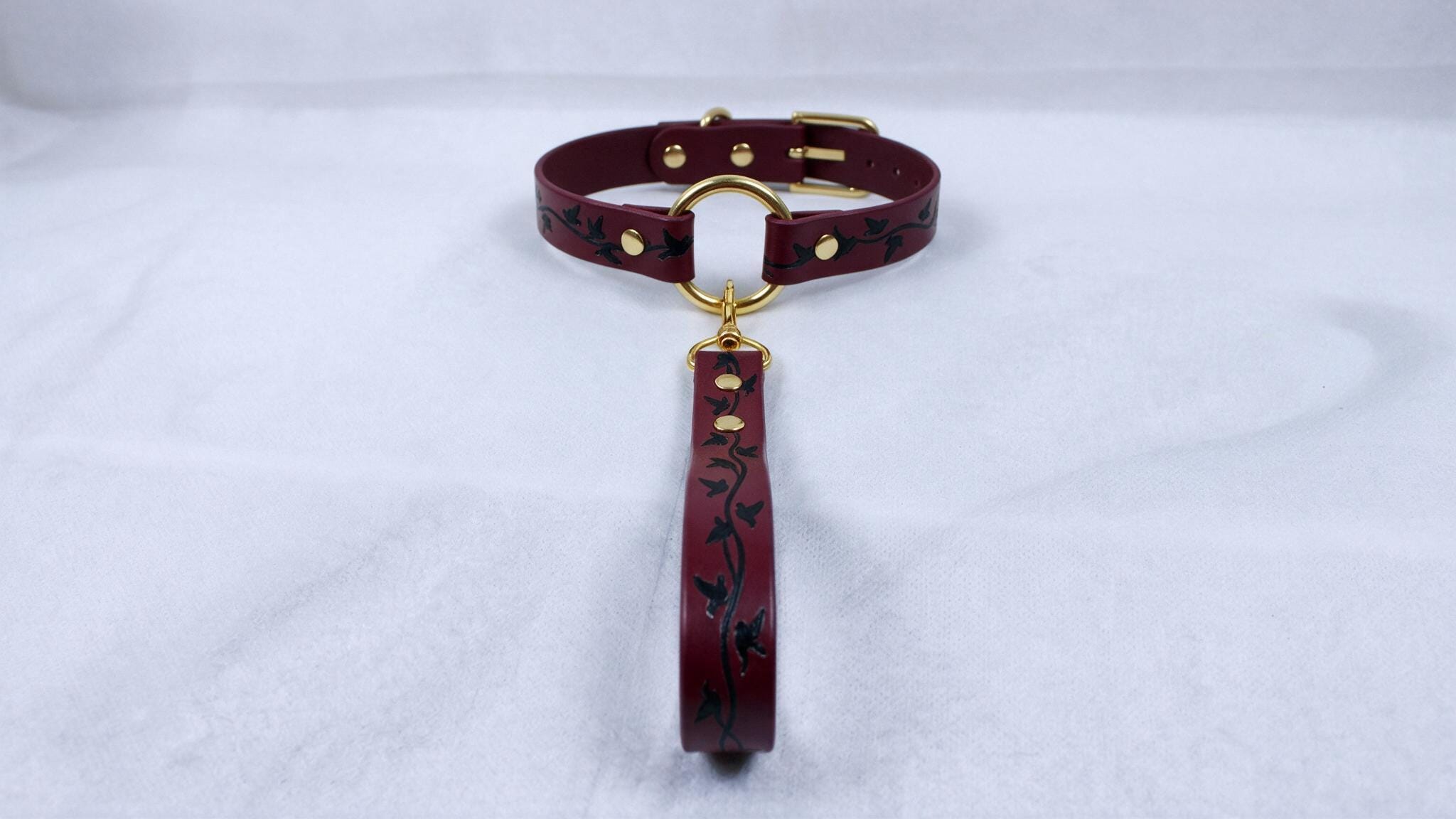 A burgundy leather collar with a gold-colored buckle and a dangling leash attached to it, resting on a white surface.