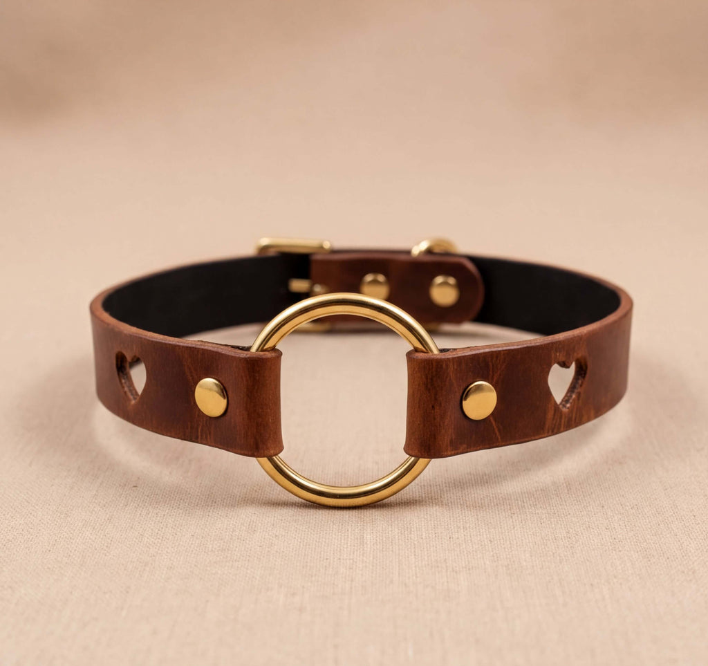 A brown leather choker collar with a gold-colored circular loop and two heart-shaped holes on the sides.