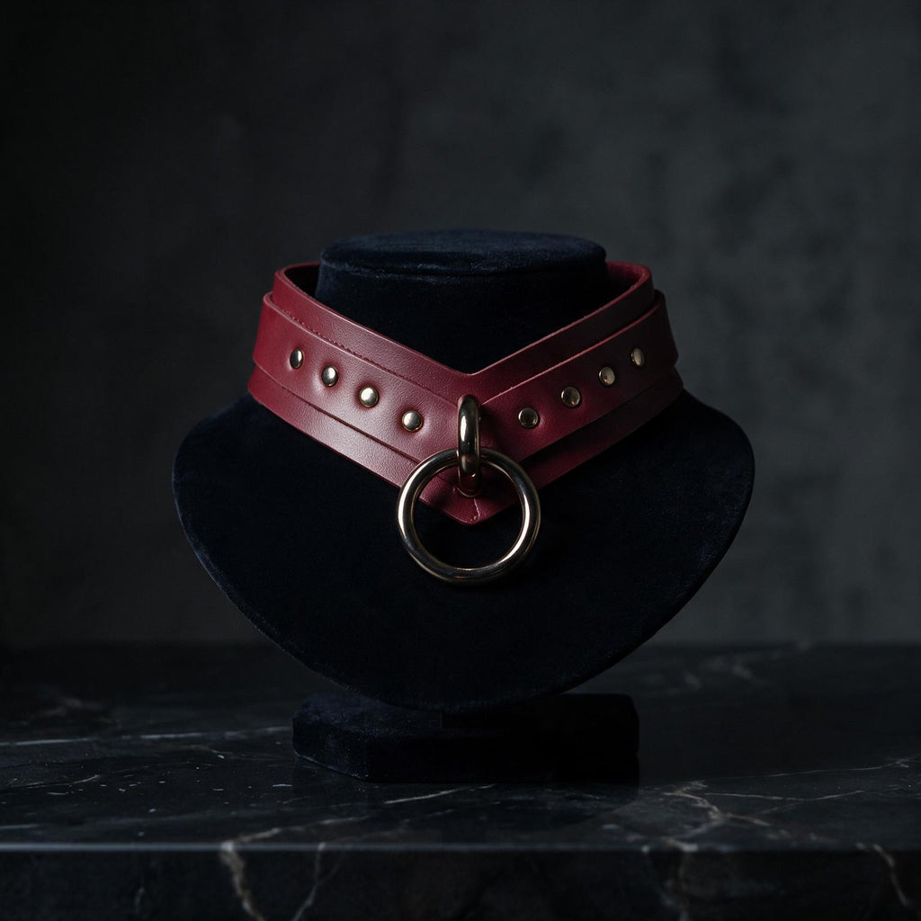A red leather choker necklace with a metal ring and studs is displayed on a black velvet stand against a dark background.