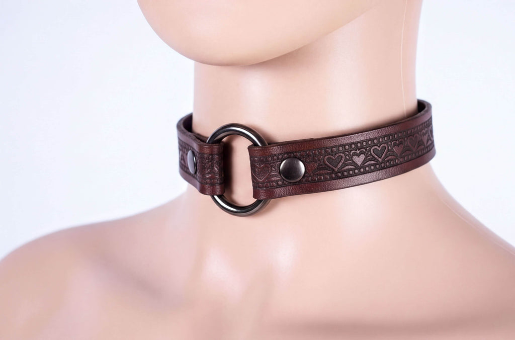 A close-up image of a mannequin wearing a brown leather choker necklace with a circular metal ring and decorative heart patterns.