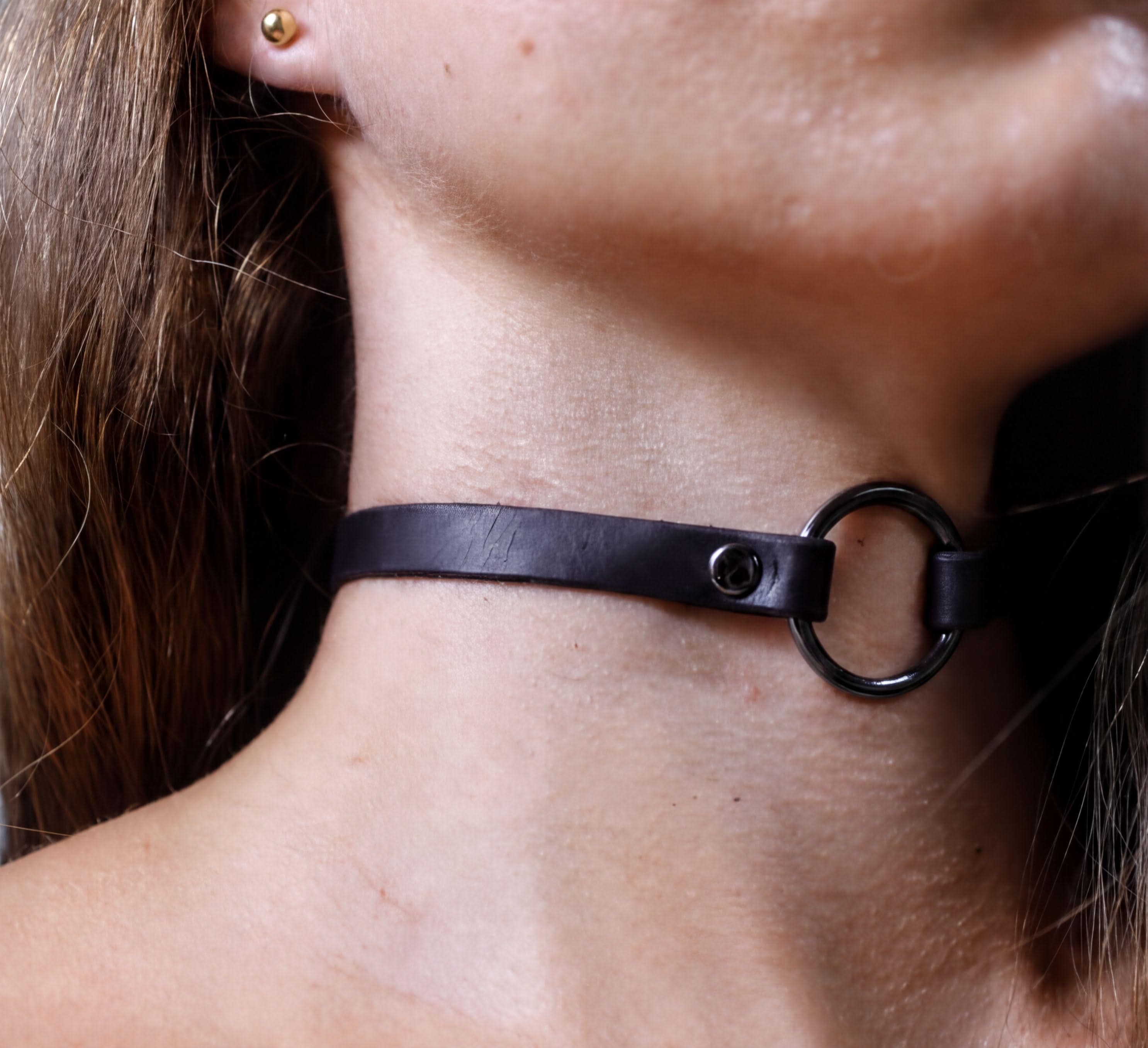 A close-up view of a person's neck and chin area, featuring a black leather choker necklace with a metal ring.