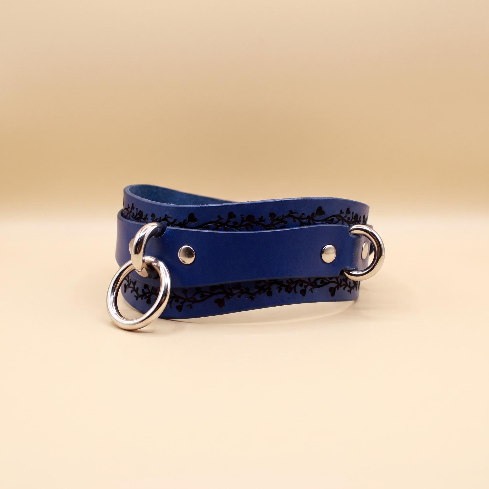 A blue leather strap with metal rings and a decorative pattern on it, placed on a beige background.
