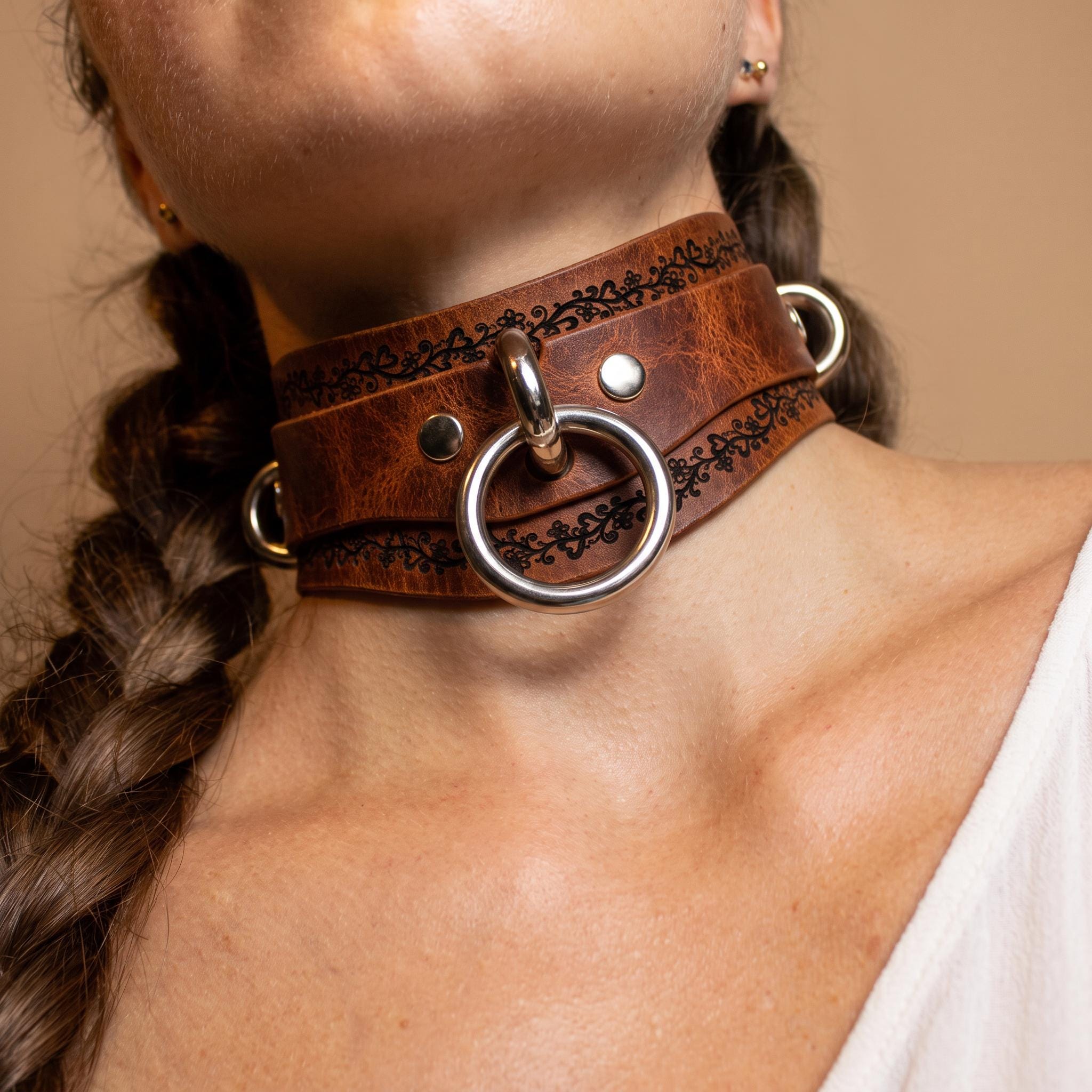 A close-up of a woman's neck wearing a brown leather choker necklace with metal rings and a metal ring.