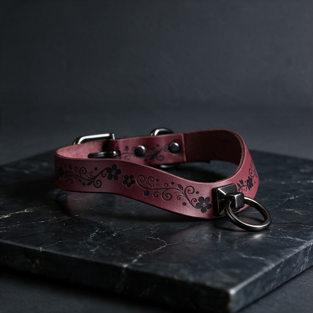 A burgundy leather choker necklace with a floral design and metal ring is displayed on a dark surface.