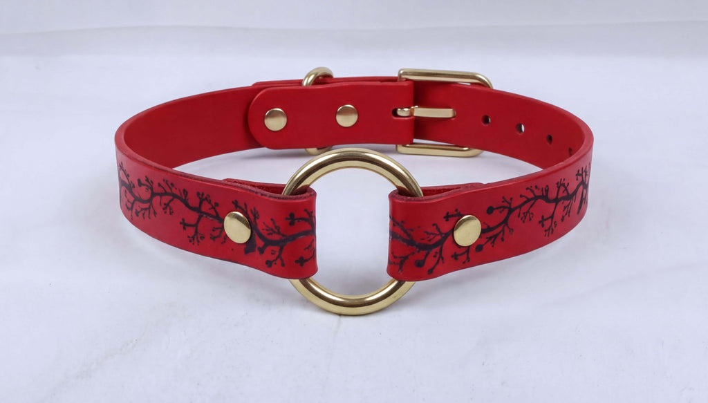 A red leather choker collar with a gold ring and a black branch design.