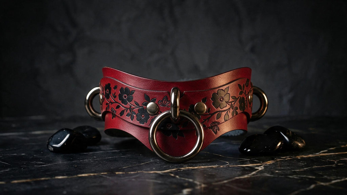 A red leather choker necklace with black floral designs and metal rings is displayed on a black marble surface, accompanied by black stones.