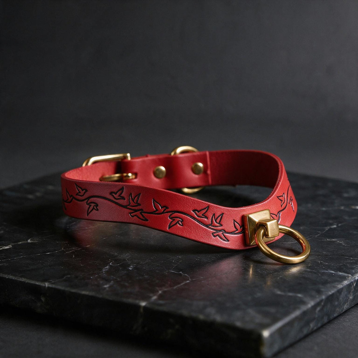 A red leather choker necklace with a gold-colored metal ring and a leaf-like design on the collar is displayed on a black marble surface.