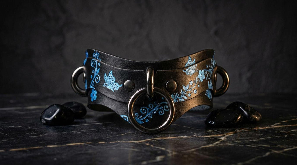 A black leather choker necklace with blue floral designs and metal rings is displayed on a dark surface, accompanied by black stones.