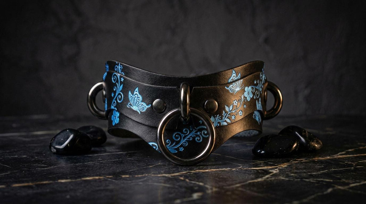 A black leather choker necklace with blue floral designs and metal rings is displayed on a dark surface, accompanied by black stones.
