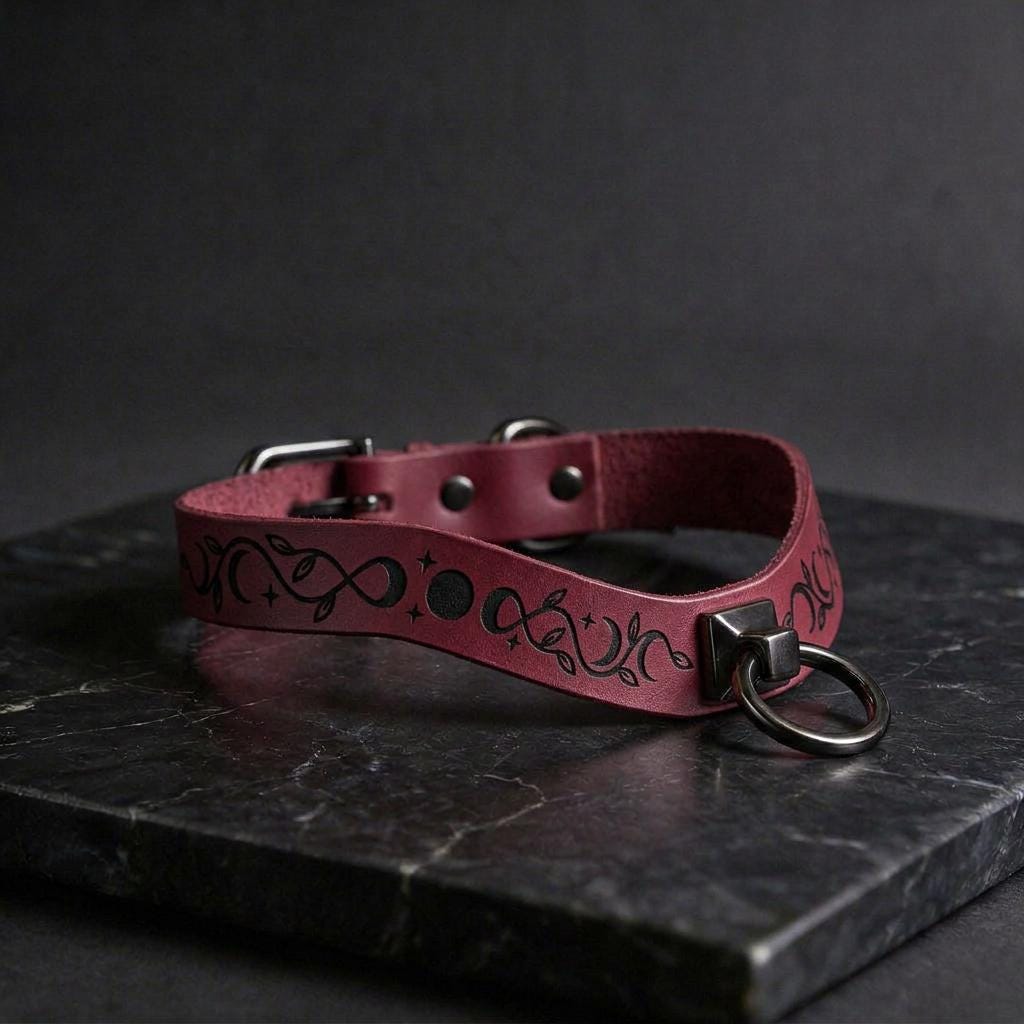 A burgundy leather choker collar with a metal ring and a decorative pattern on the strap is displayed on a dark surface.