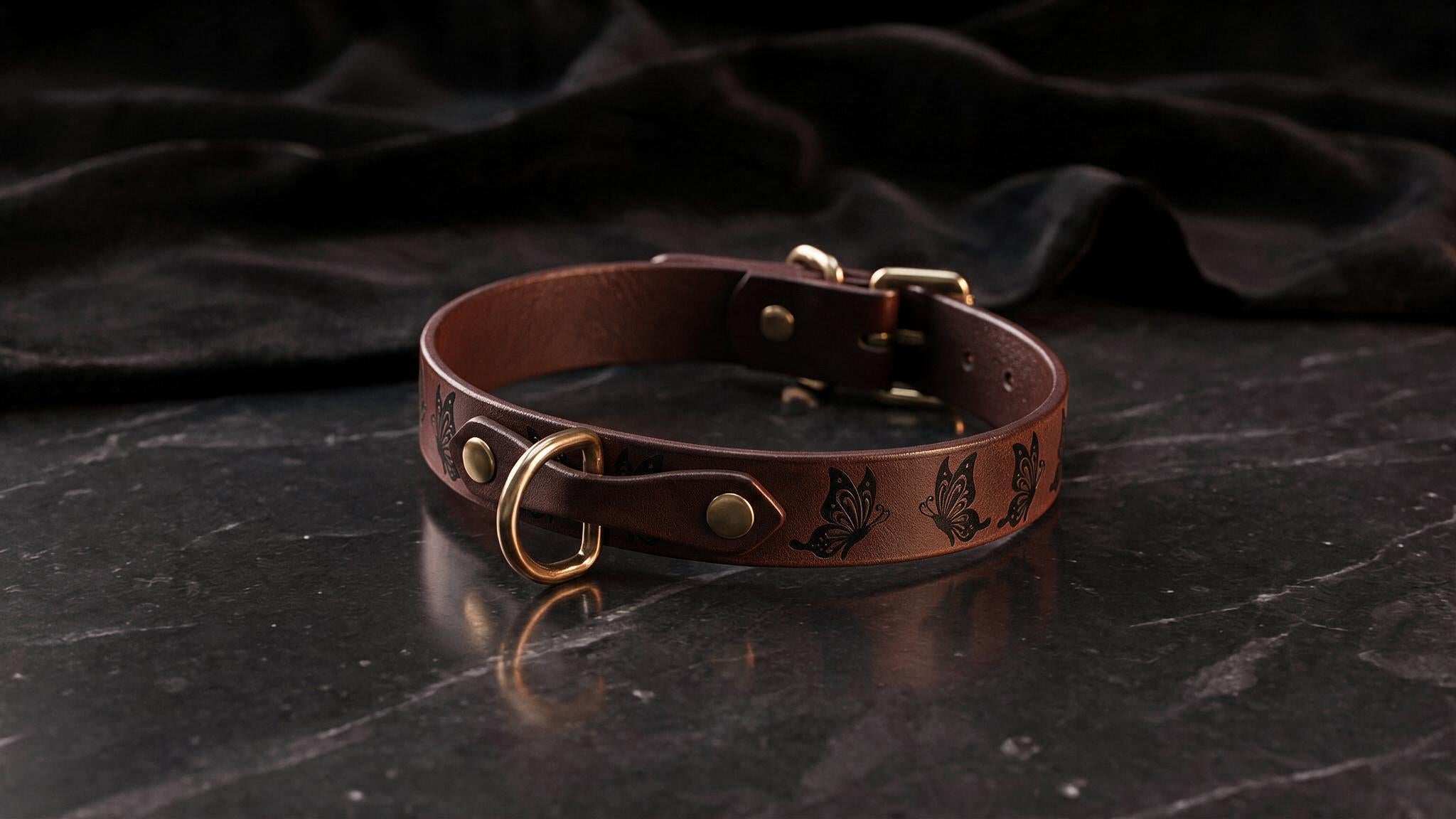A brown leather collar with a gold buckle and butterfly design, resting on a dark surface.