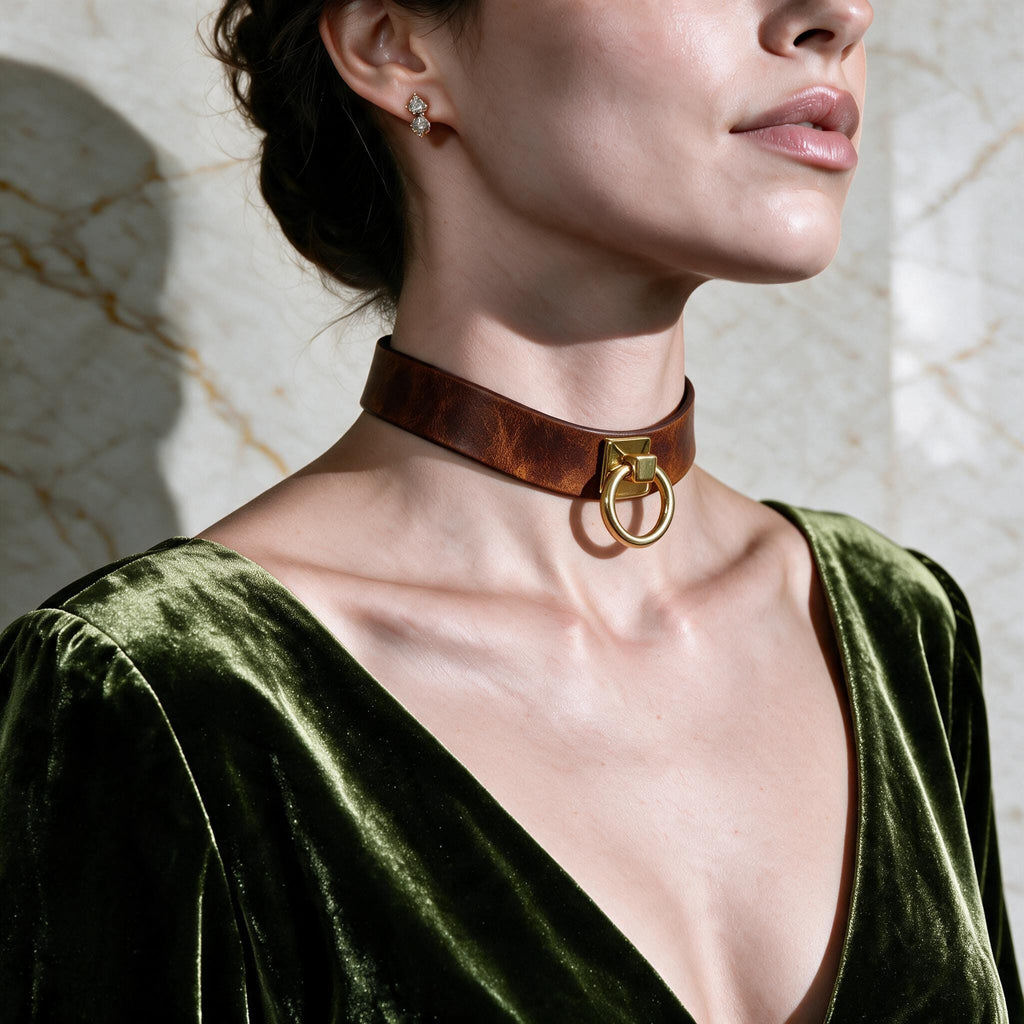 A woman wearing a brown choker necklace with a gold ring on it, and a green velvet dress, posing against a marble wall.