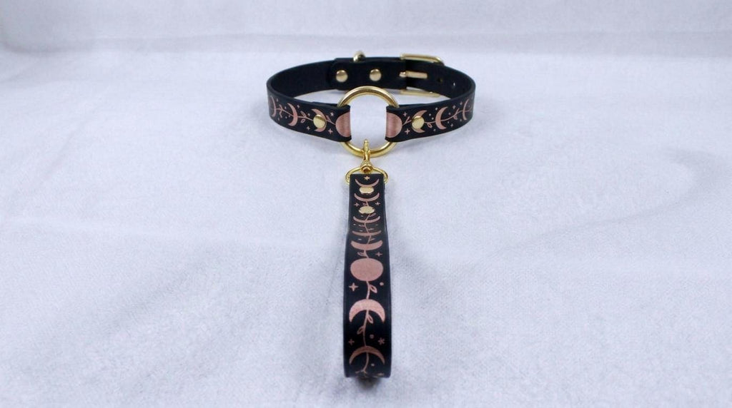 A black collar with a gold-colored buckle and a decorative pattern on the strap is displayed against a white background.