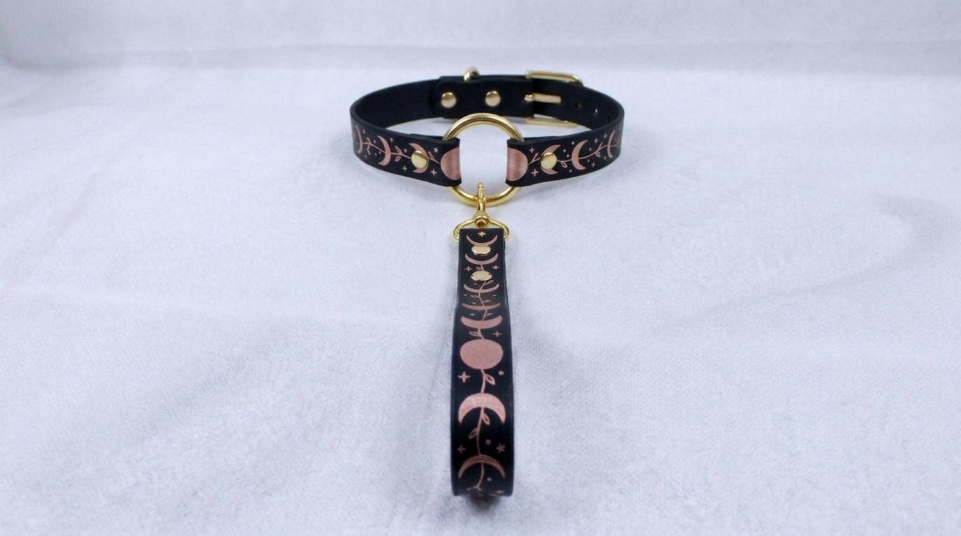 A black collar with a gold-colored buckle and a decorative pattern on the strap is displayed against a white background.