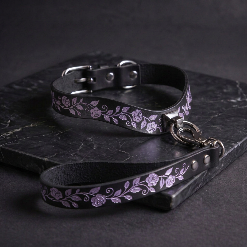 A black leather collar with a floral pattern and a silver metal buckle is displayed on a dark surface.