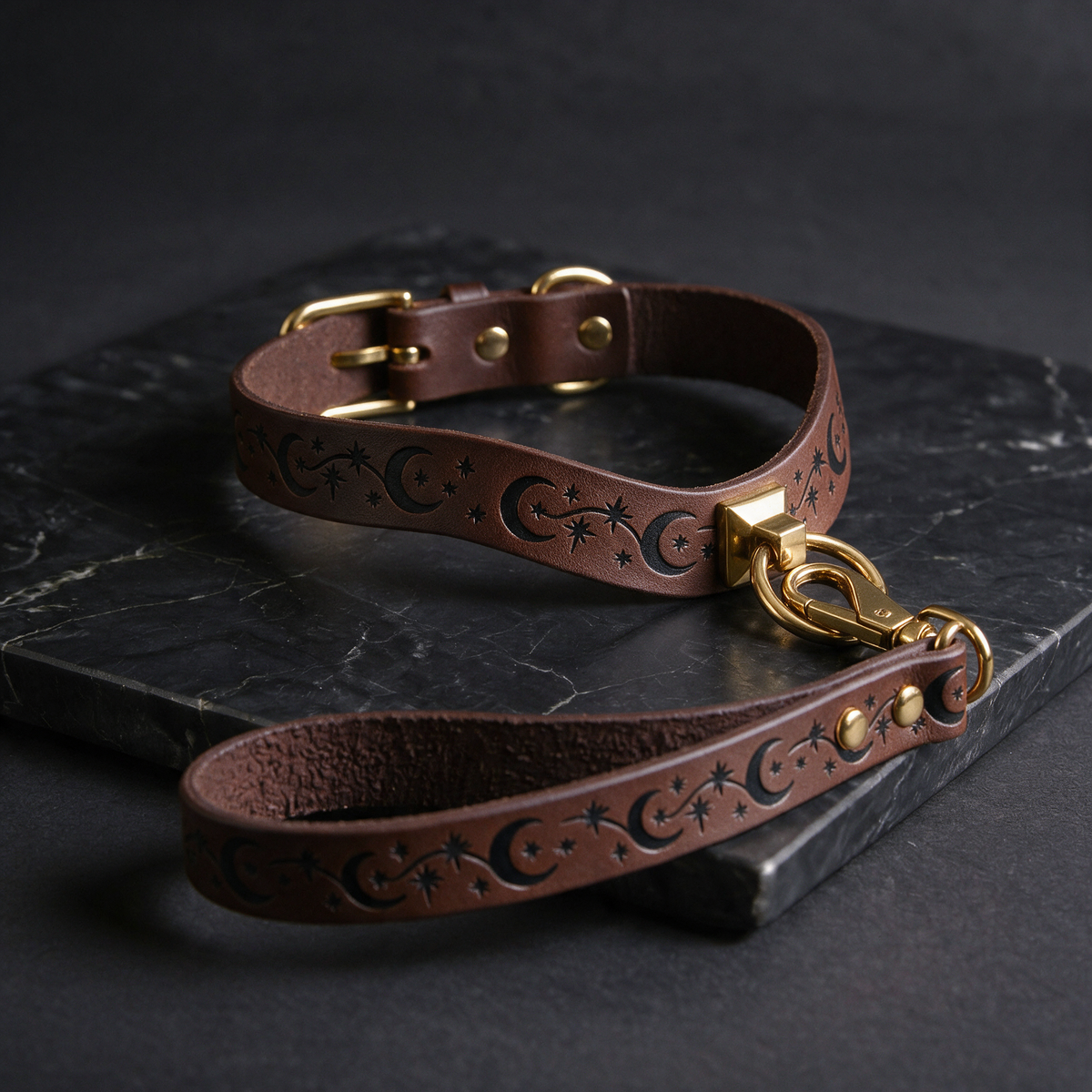 A brown leather dog collar with a star and crescent design, featuring a gold buckle and a matching gold-colored leash attached to it, resting on a dark surface.