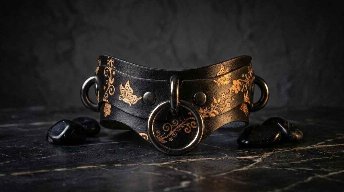 A black leather choker necklace with gold floral designs and metal rings is displayed on a dark surface, accompanied by black stones.